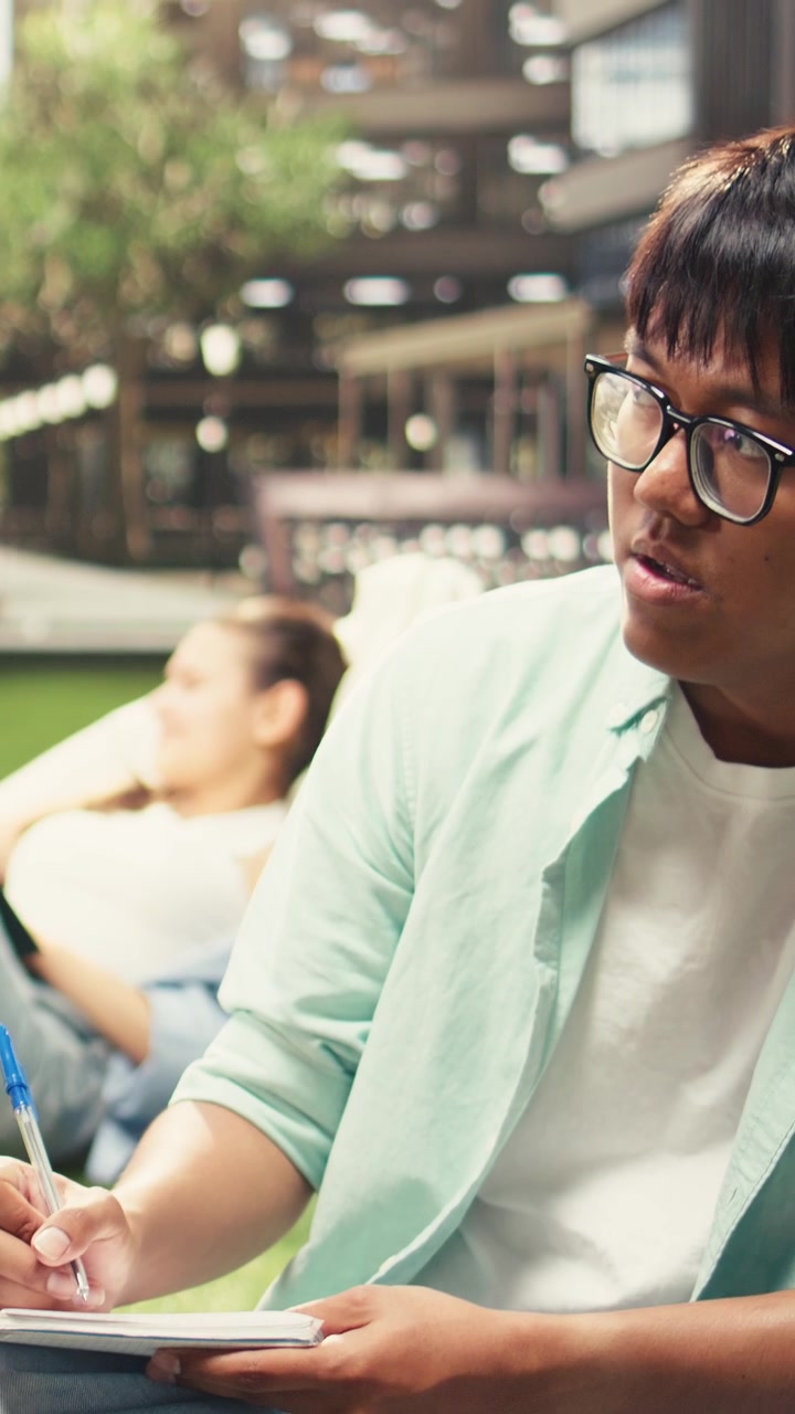 Vertical Video Classmates in study session sitting on grass with laptop placed on stacked books