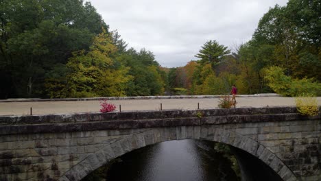 Antena-De-Un-Joven-Camina-Por-Un-Camino-De-Grava-En-Un-Puente-Sobre-Un-Río-En-Otoño