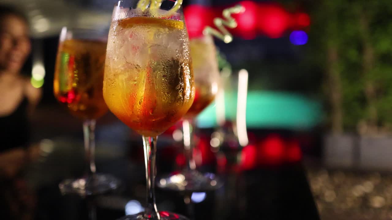 Multiple glasses of orange cocktails with ice and garnish on a reflective table, illuminated by vibrant, colorful evening lights in an outdoor garden setting. Subtle camera push-in