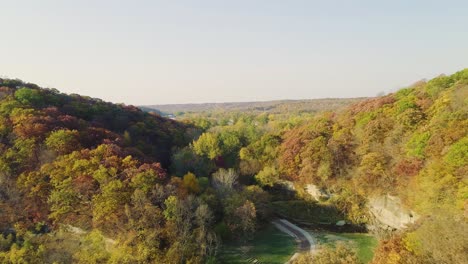 Flyover-of-autumn-trees-and-hills-at-Ledges-State-Park-at-sunrise,-revealing-a-road-winding-between