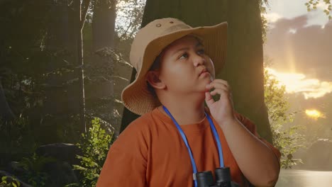 niño asiático con un sombrero y binoculares pensando en algo y mirando a su alrededor mientras explora la naturaleza forestal. niño investigador, concepto de aventura de turismo de viaje, de cerca