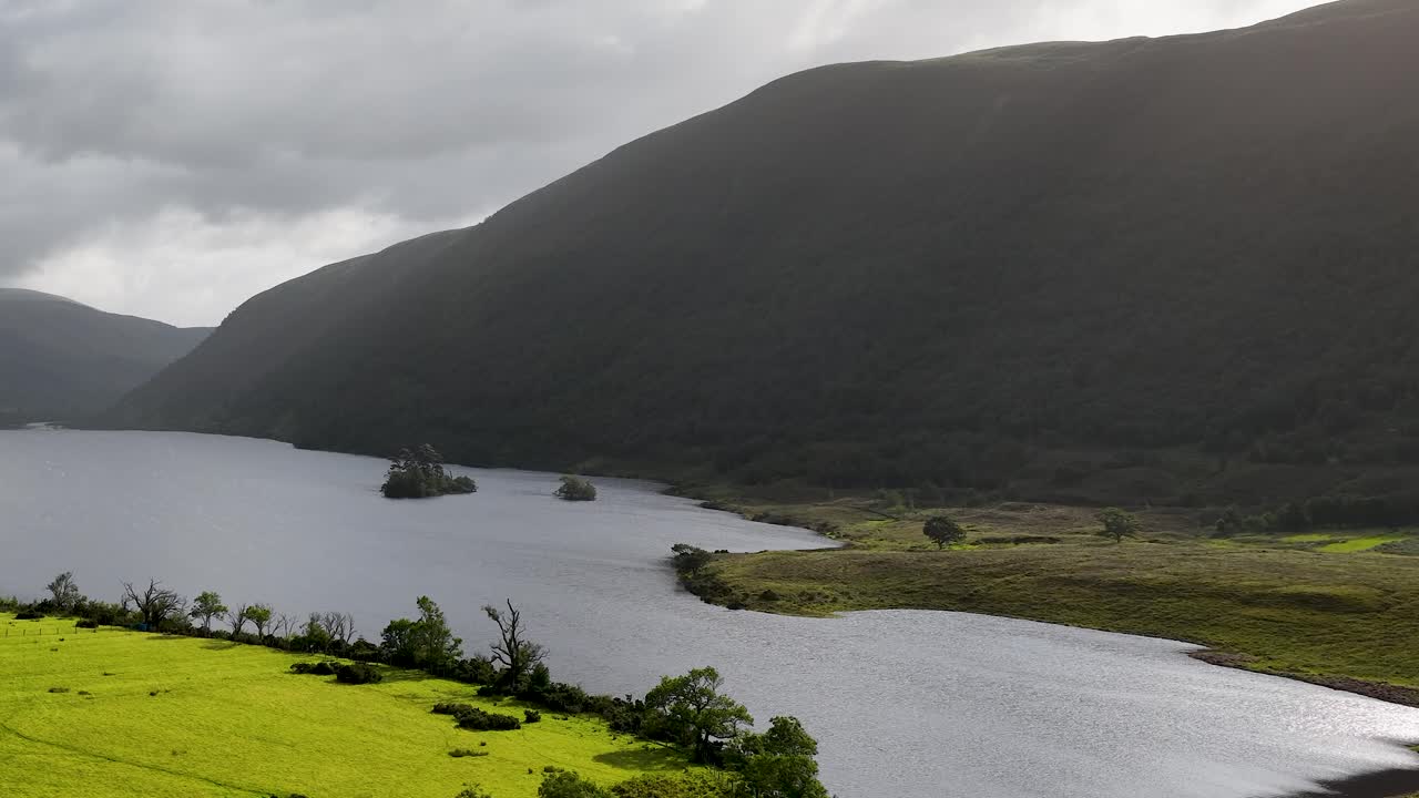 Wide shot pans over serene lake, green meadows, and mountains under overcast natural daylight