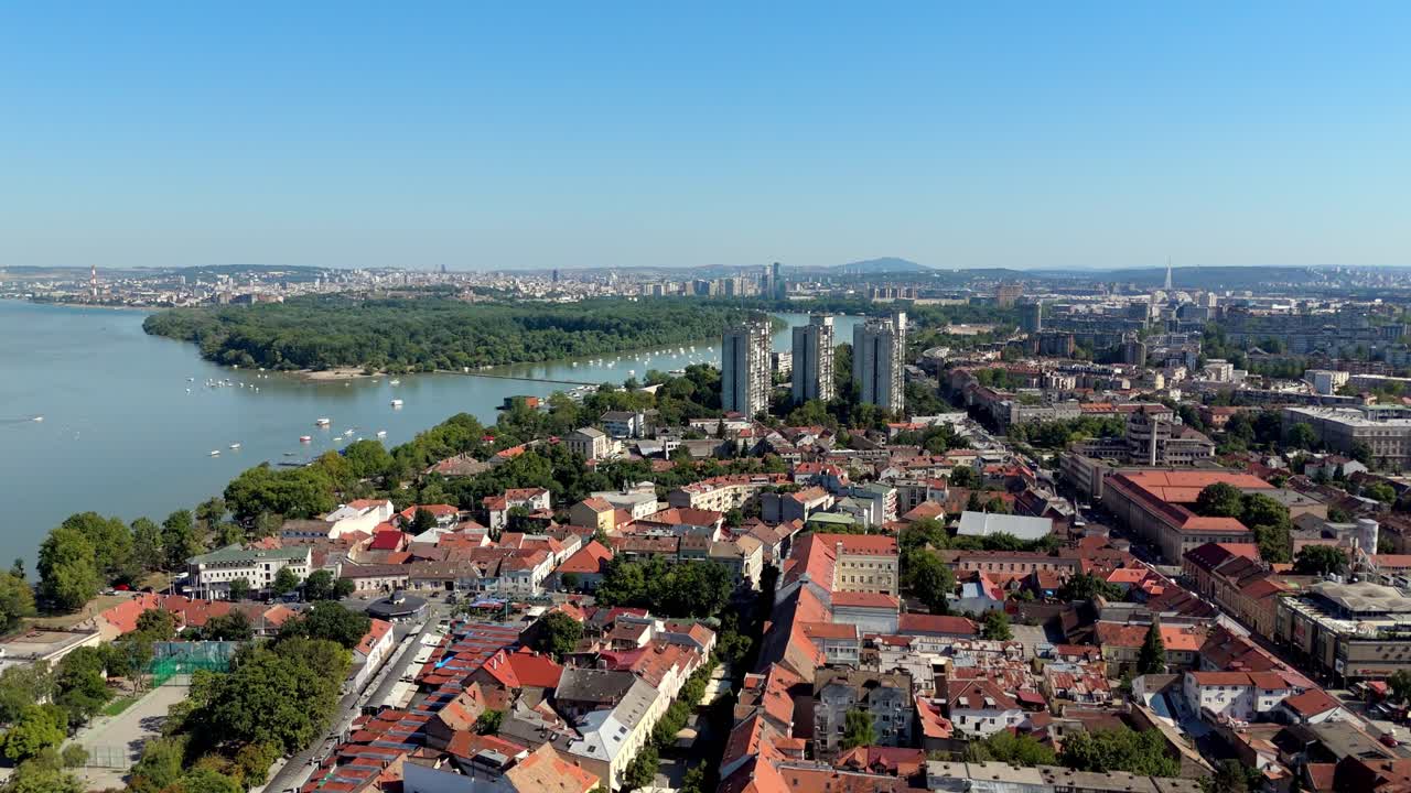 Aerial drone view of Belgrade skyline, Serbia, overlooking the confluence of the Sava and Danube rivers, with modern towers, red rooftops, and lush greenery under clear blue sky