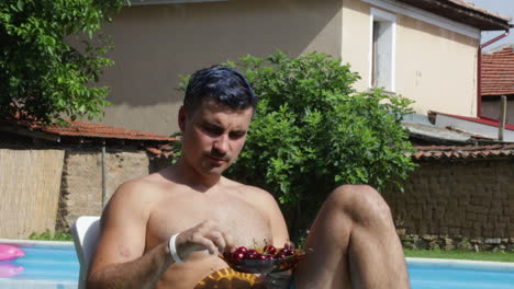 young man sits on poolside bed and eats cherries, front view