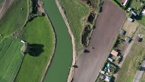 Toma-Aérea-De-Arriba-Hacia-Abajo-De-Un-Río-Con-Curvas-Que-Corre-Entre-Los-Campos,-Granjas-Y-Jardines-De-Un-Pueblo-Agrícola-Rural