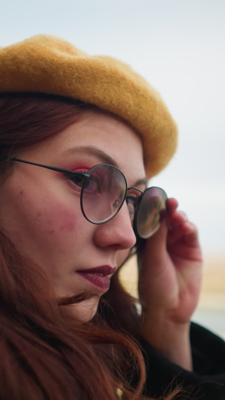 primer plano de una mujer alegre con una boina amarilla y gafas de sol, sus labios pintados curvados en una sonrisa, con un telón de fondo borroso de árboles de otoño, un río y una estructura de madera