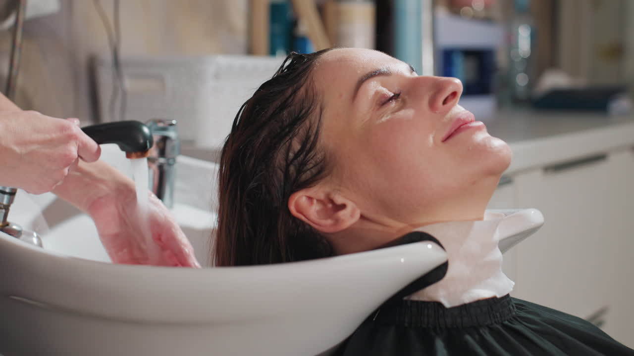 백인 고객이 편안하게 기대어 앉아 두피 관리를 받고 있으며, 스타일리스트가 따뜻한 물로 마사지하고 있습니다. 헤어라인을 따라 부드럽게 압력을 가하고, 목 아래에는 수건을 받쳐 줍니다. 편안한 분위기의 테라피 살롱입니다.