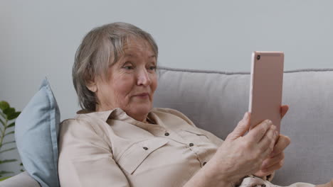 happy senior woman having a videocall via tablet while lying on sofa at home 2