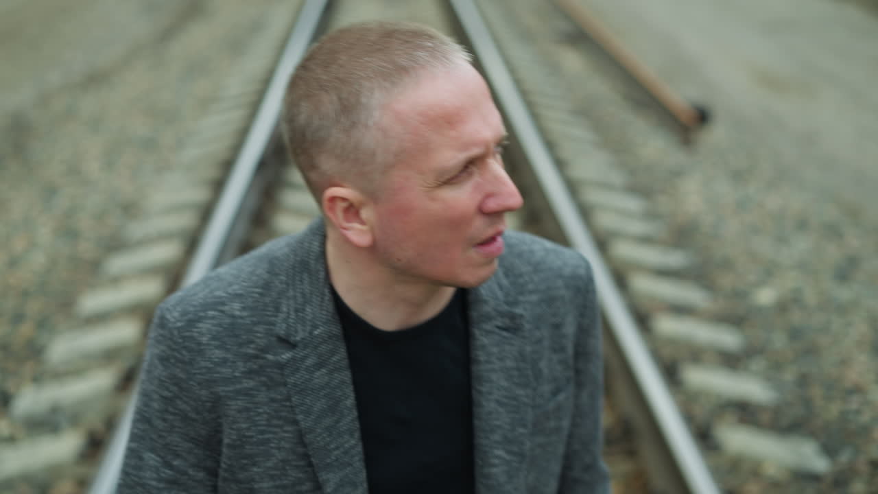 близкий вид пожилого белого человека в сером костюме и черной рубашке поло, отворачивающегося от камеры, идущего по железной дороге с камнями, с размытым видом деревьев и электрических столбов с обеих сторон