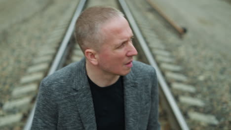 a close view of an aged white man wearing a grey suit and black polo shirt, looking away from the camera, walking on a railway with stones, with blur view of trees and electric poles on either side