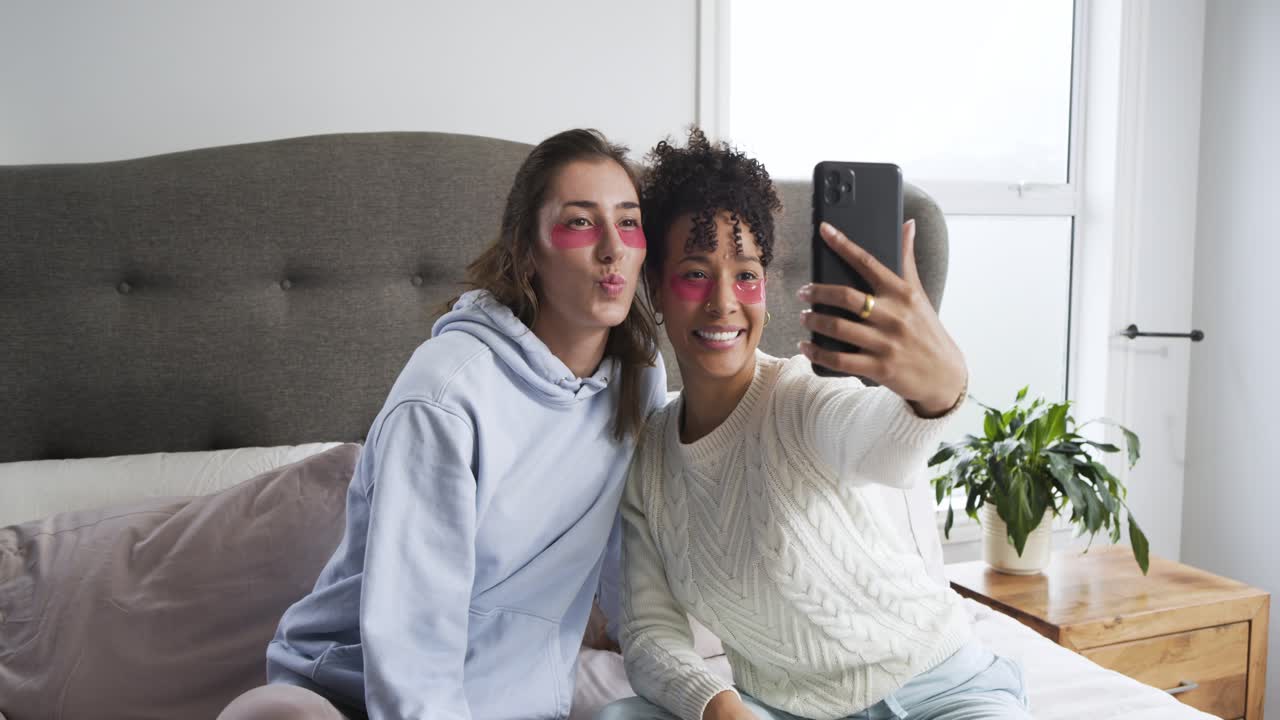 Diverse amiche che si applicano patch occhi sul letto prima di posare e rivedere le immagini dello smartphone.
