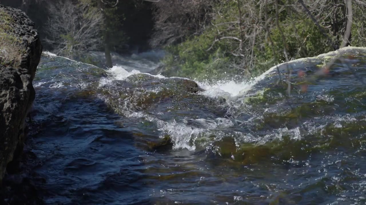 Una vista dall'alto cattura l'acqua del fiume che scorre rapidamente sulla cresta di un'enorme cascata calcarea. Schiuma bianca e correnti scure creano motivi ad alto contrasto
