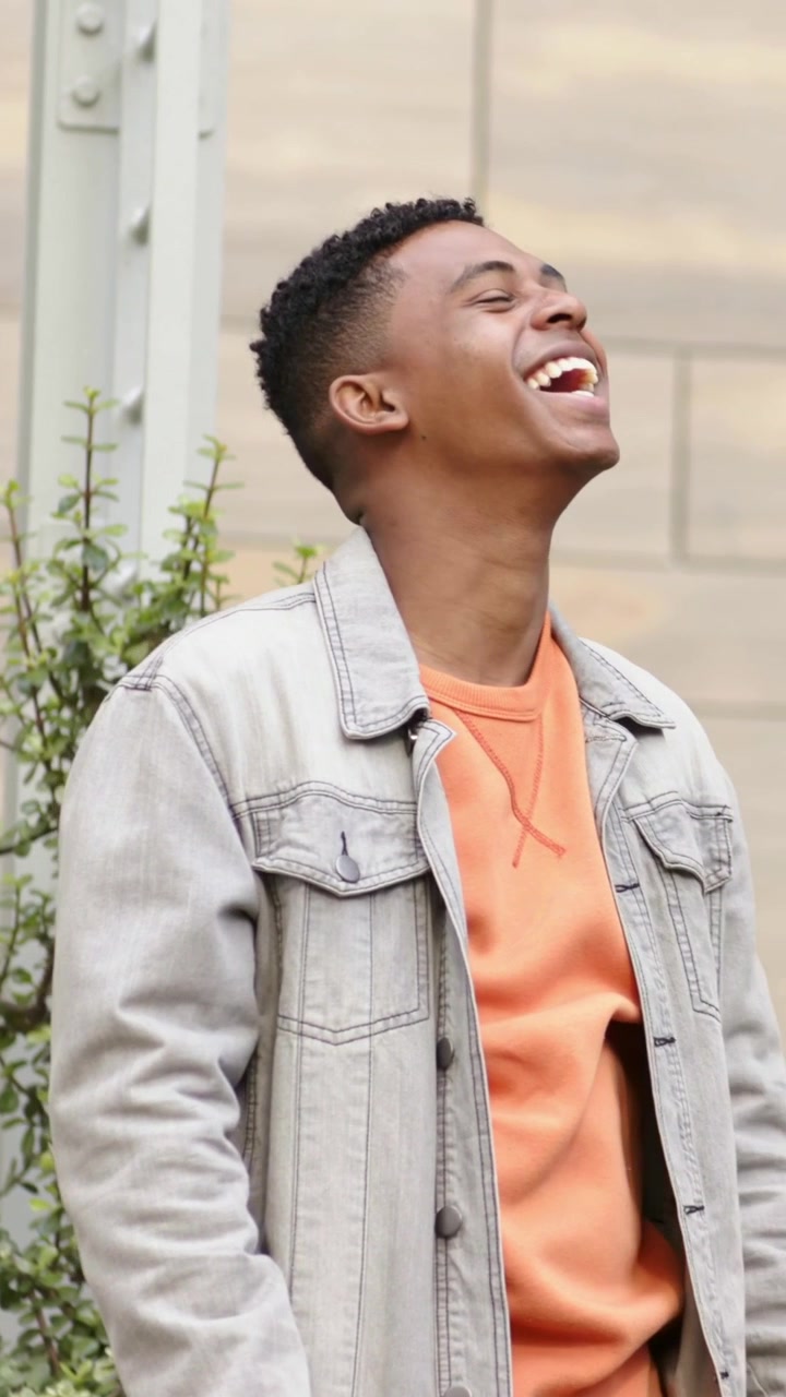 Vertical video: Laughing man in orange sweatshirt talking off-camera by tiled wall, grabbing phone