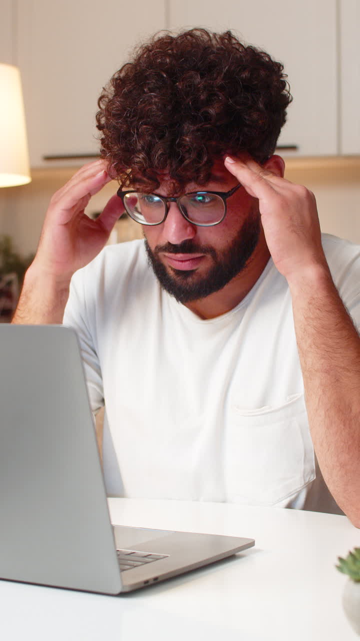 Arabian man holds head from screen strain takes break then resumes laptop work evening indoors
