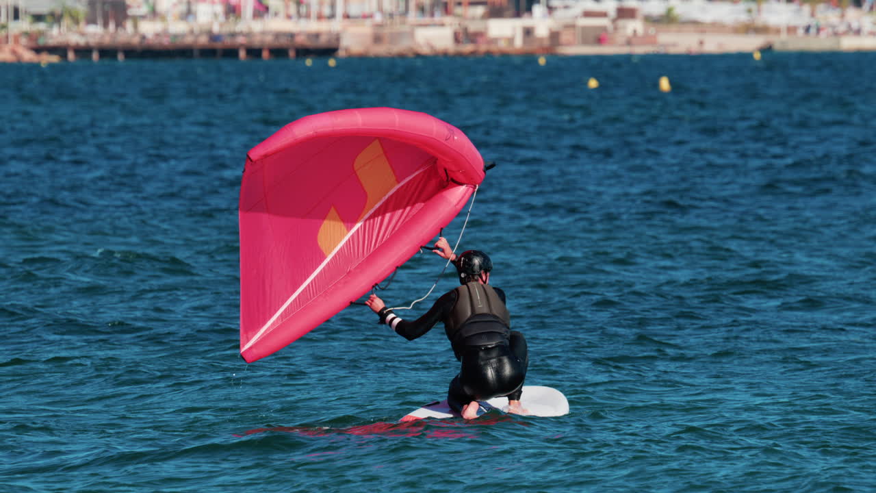 A man in a wetsuit practices wing foiling with a bright pink sail on the blue Mediterranean waters of Cannes, France