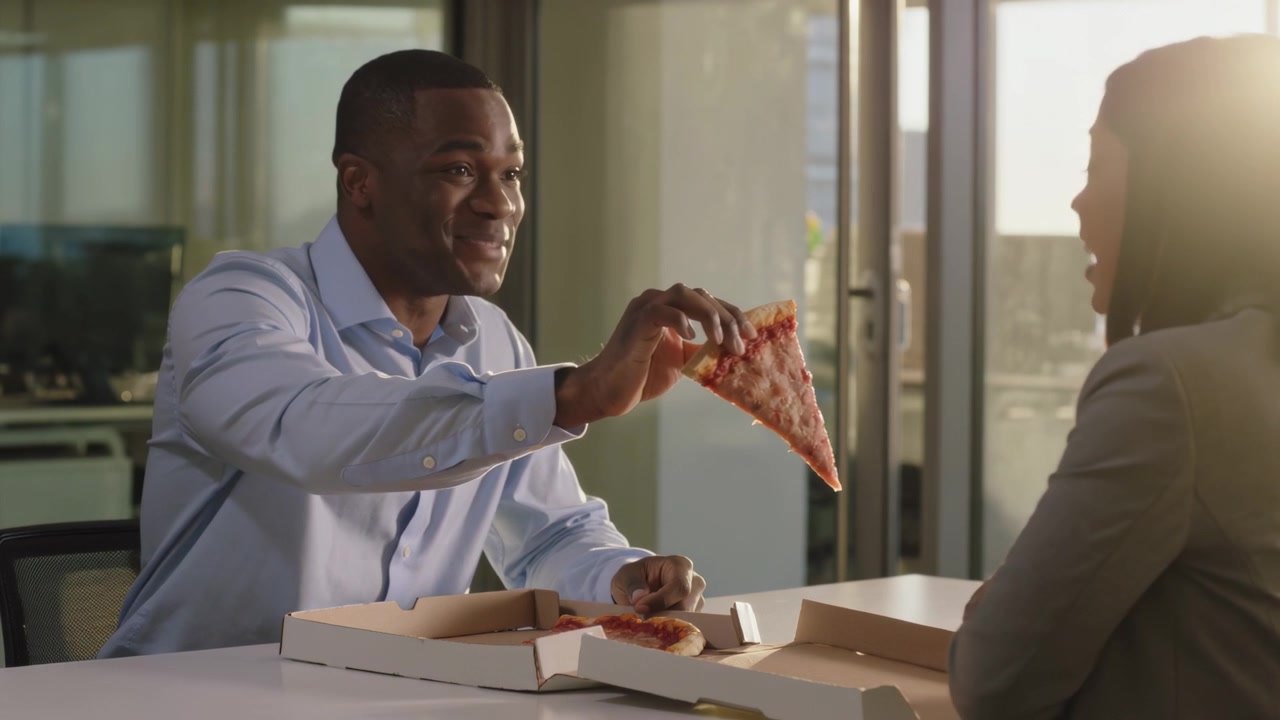 Opening pizza box, man in blue shirt offering slice to colleague in gray suit during meeting