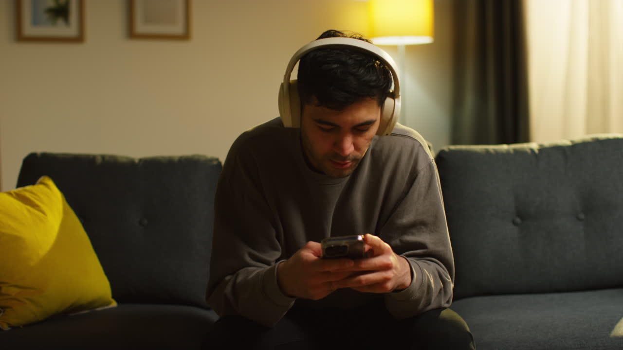 joven que pasa la noche en casa sentado en el sofá con auriculares inalámbricos transmitiendo música o viendo contenido desde el teléfono móvil