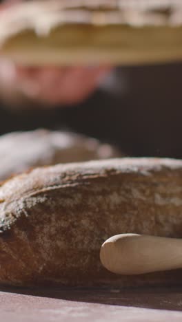 Vertical-Video-Low-Key-Shot-Of-Person-Putting-Freshly-Baked-Loaf-Of-Bread-Onto-Work-Surface