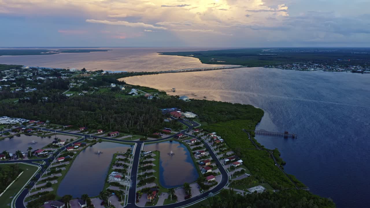 A peaceful coastal community sits along reflective bay waters, where curving streets, small lakes, and lush greenery meet the soft golden glow of sunset over the horizon