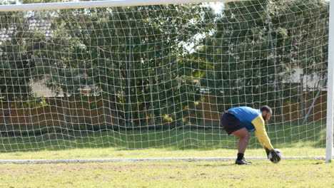 goalkeeper in soccer uniform bending to catch ball in front of goal net