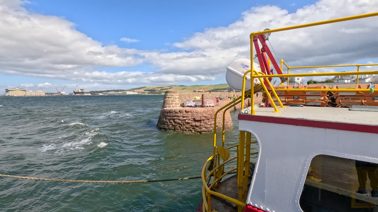 A ferry moves away from a stone harbor wall into open water under partly cloudy skies, with dynamic waves and bright daylight highlighting the coastal scene