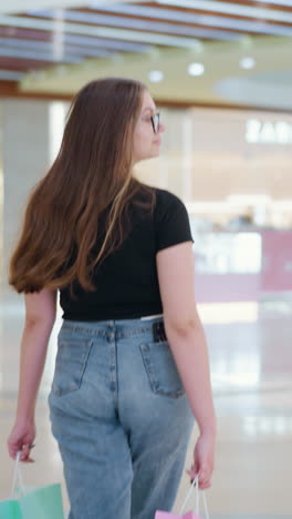 쇼핑몰에서 쇼핑백을 들고, 돌아서서 호기심을 가지고 주위를 둘러보며, 활기찬 쇼핑 센터를 걸어가면서 머리카락을 과시하고, 다른 쇼핑객들이 지나가고 있습니다.
