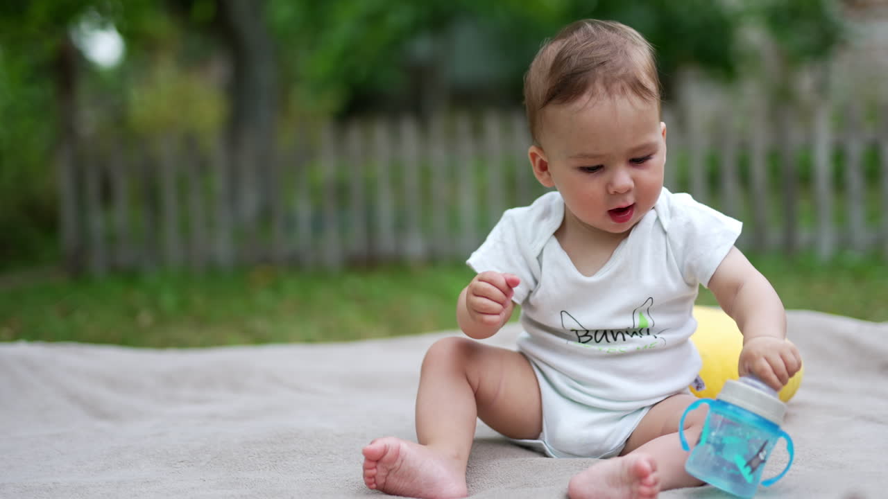 Tiny toddler sitting outdoors playing with his bottle of water. Lovely baby tries to change position to crawl ahead. Blurred backdrop.