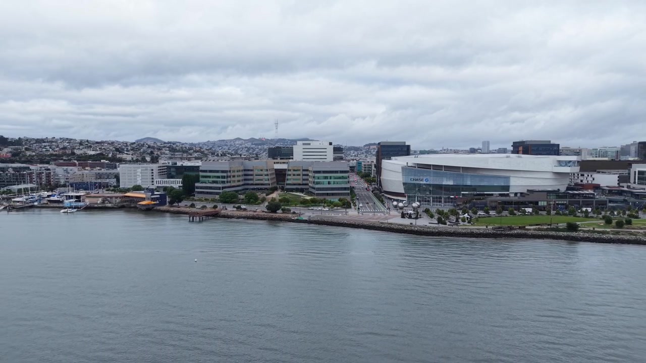 Ripresa drone che cattura il moderno lungomare di Mission Bay, l'arena Chase Center e il paesaggio urbano di San Francisco sotto cieli coperti, ripresa dall'alto con movimento dinamico del drone. Ripresa drone in allontanamento