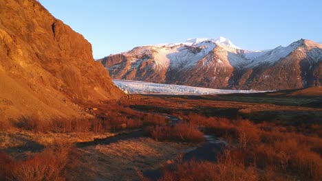 Stunning-aerial-establishing-wide-shot-of-Snaefellsjokull-Glacier-in-Iceland,-empty-trail-leading-to-the-glacier,-on-a-golden-light-bathed-landscape-and-snow-capped-rugged-mountains