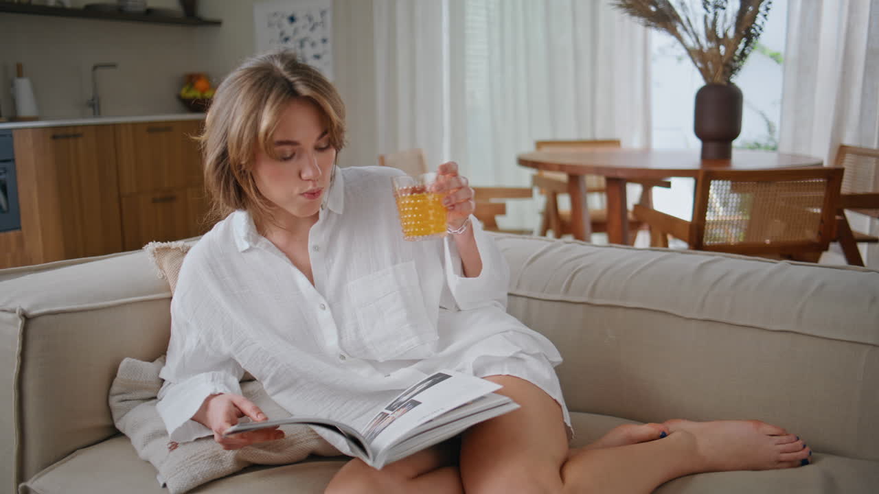 Home girl reading book sitting cozy sofa living room. Young lady drinking juice