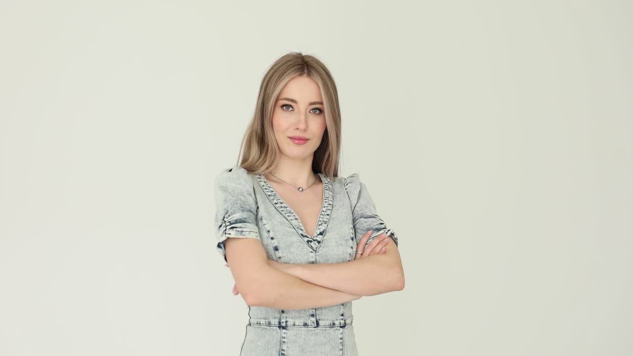 A young woman with blonde hair and a denim dress standing with crossed arms against a white background