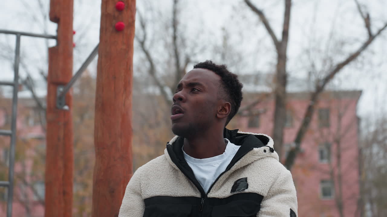 흑인 남자가 은 겨울 공기 밖에서 팔을 고 눈에 보이는 숨결, 배경에 운동 장비, 눈으로 인 땅, 잎없는 나무, 주차된 자동차 및 도시 건물