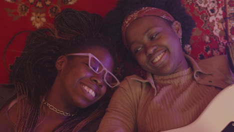 happy young girl playing guitar and talking with her friend while lying together on floor during rehearsal in recording studio