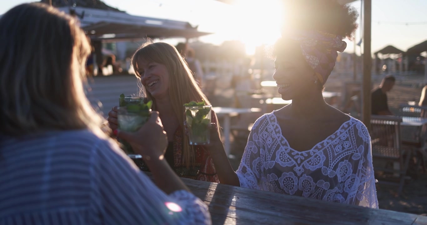 Donne felici di diverse etnie che brindano con Mojito in un bar sulla spiaggia al tramonto.