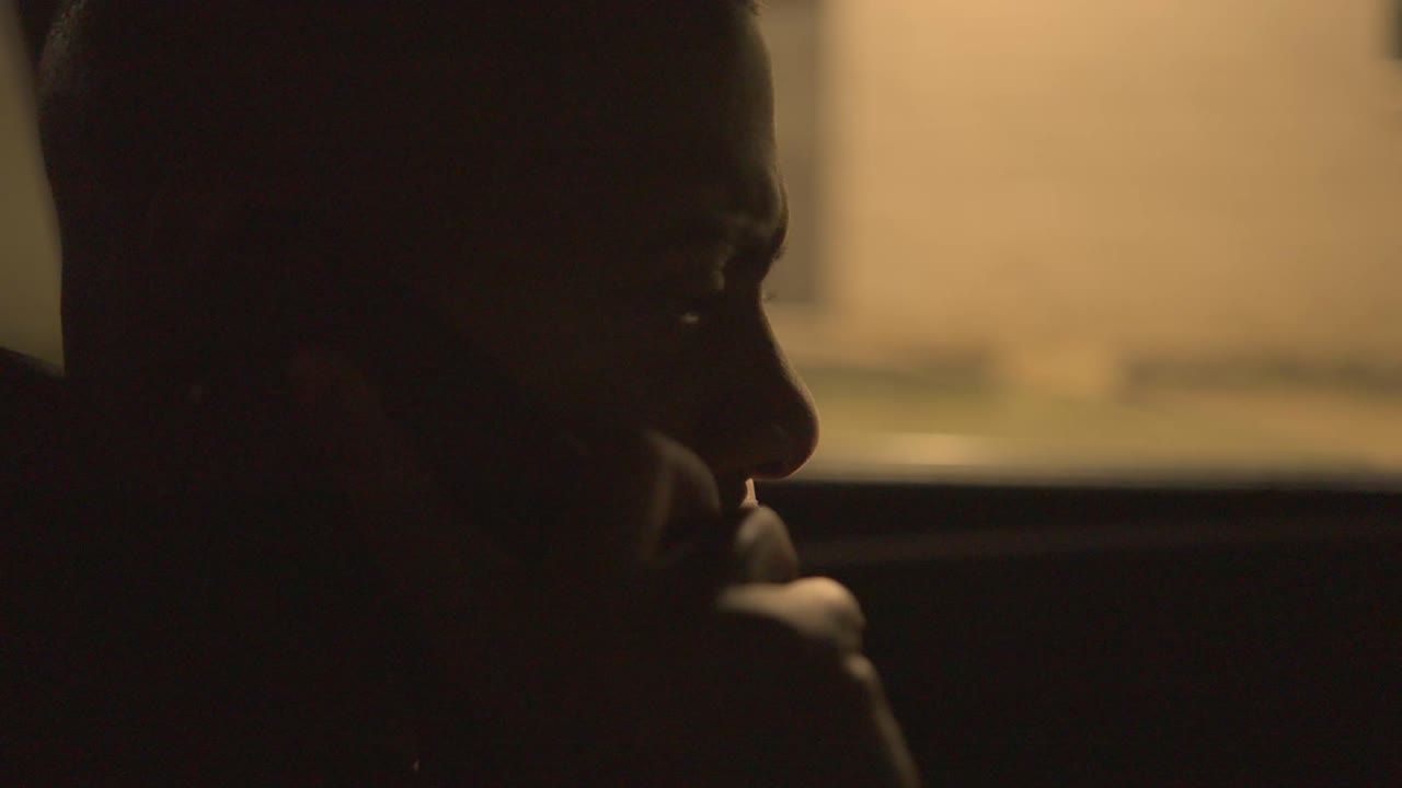 um homem negro sentado no banco do condutor de um carro fora de um armazém à noite faz um telefonema e recebe notícias perturbadoras ele está claramente angustiado e zangado