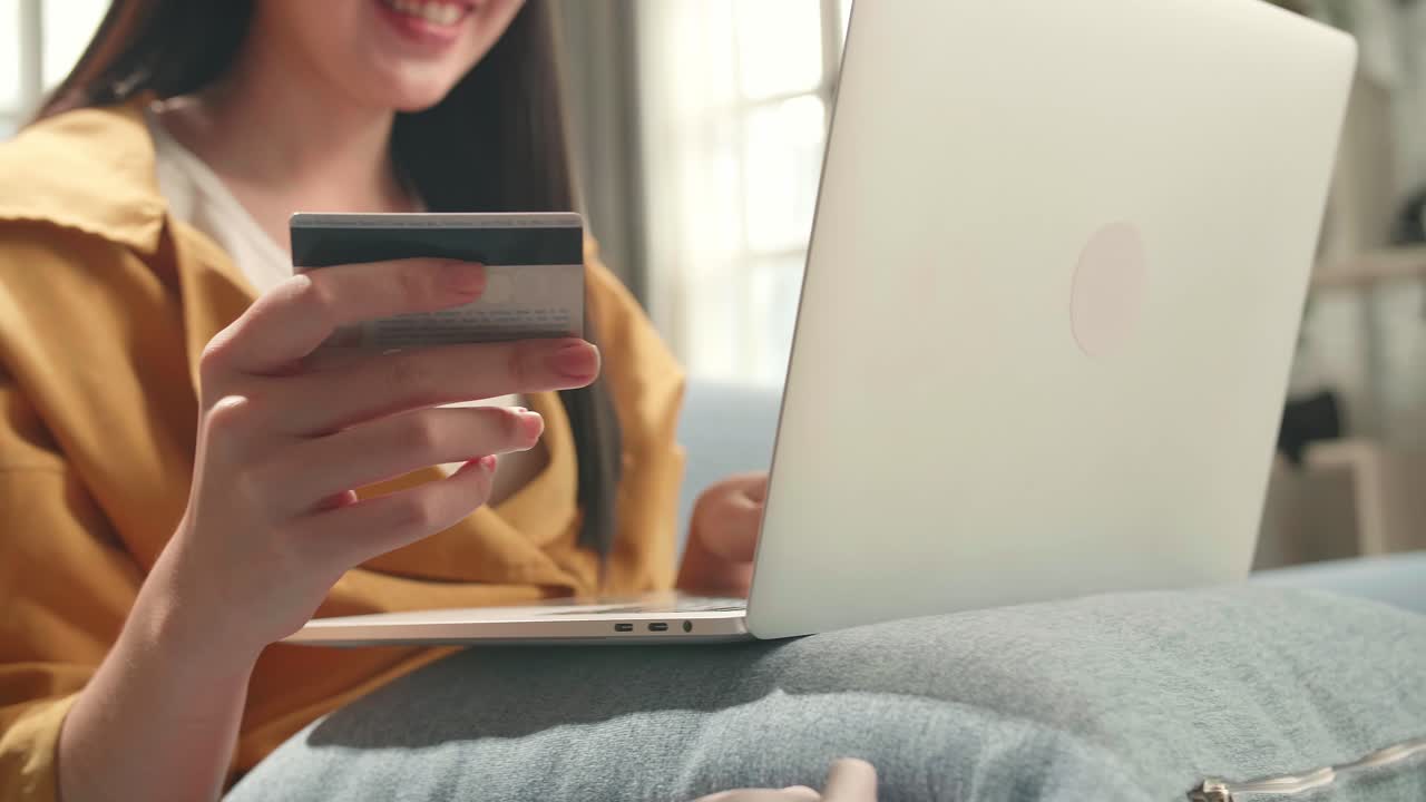 Woman Sitting On Couch In Living Room Holding Credit Card And Using Laptop Computer Shopping Online