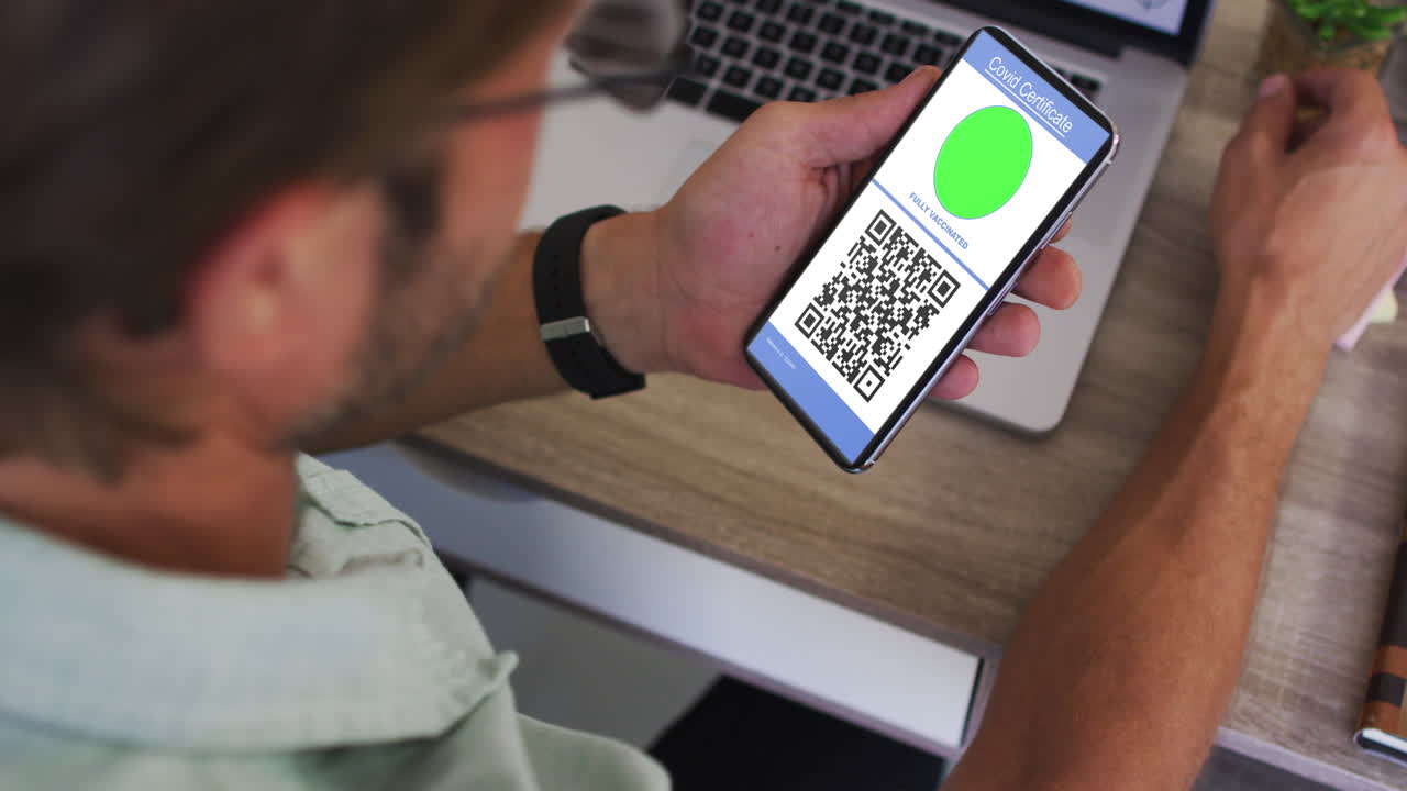 Man at desk holding smartphone with covid vaccination certificate and qr code on screen