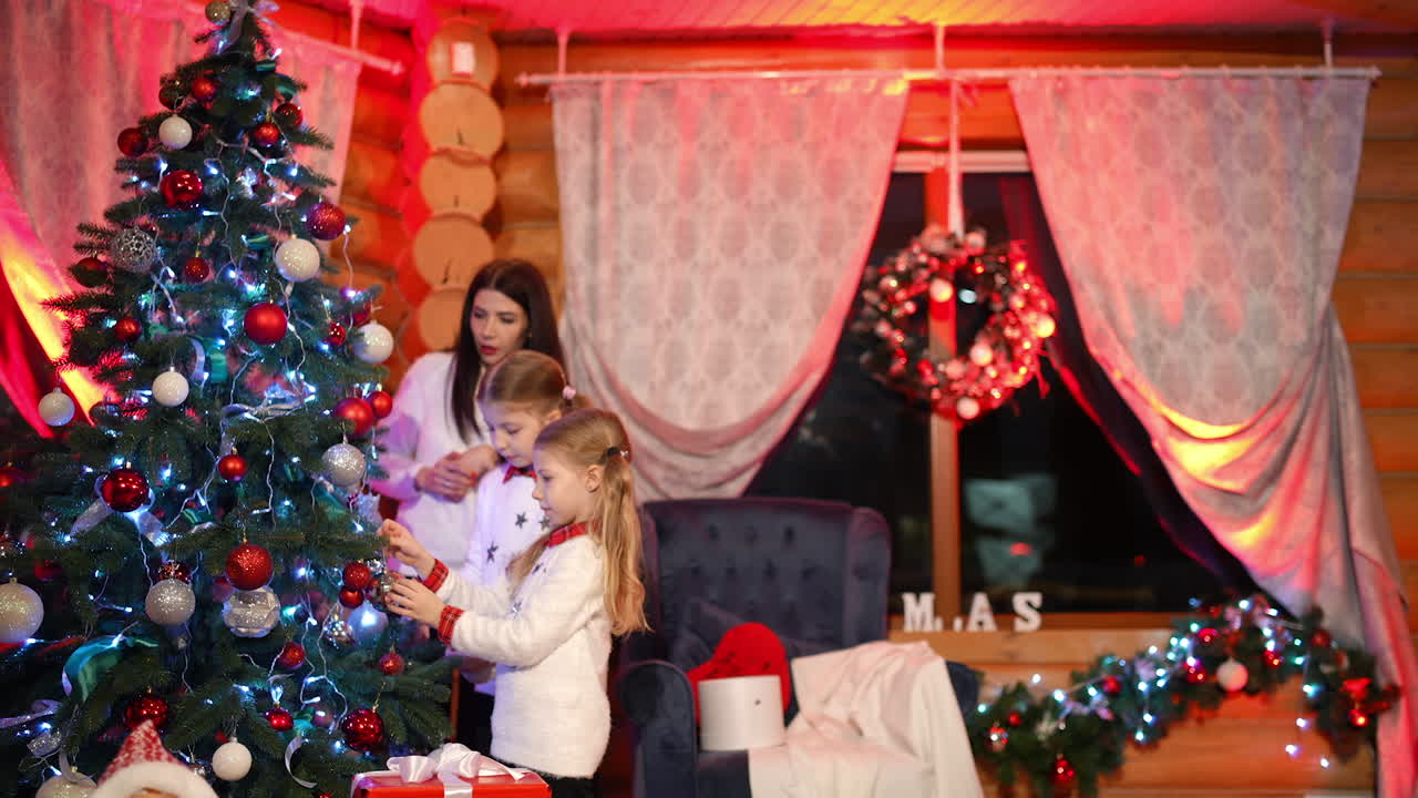 Festive atmosphere in the house before Christmas. Little girls and their mother in white sweaters decorate New Year tree together. Winter holidays and celebrations.