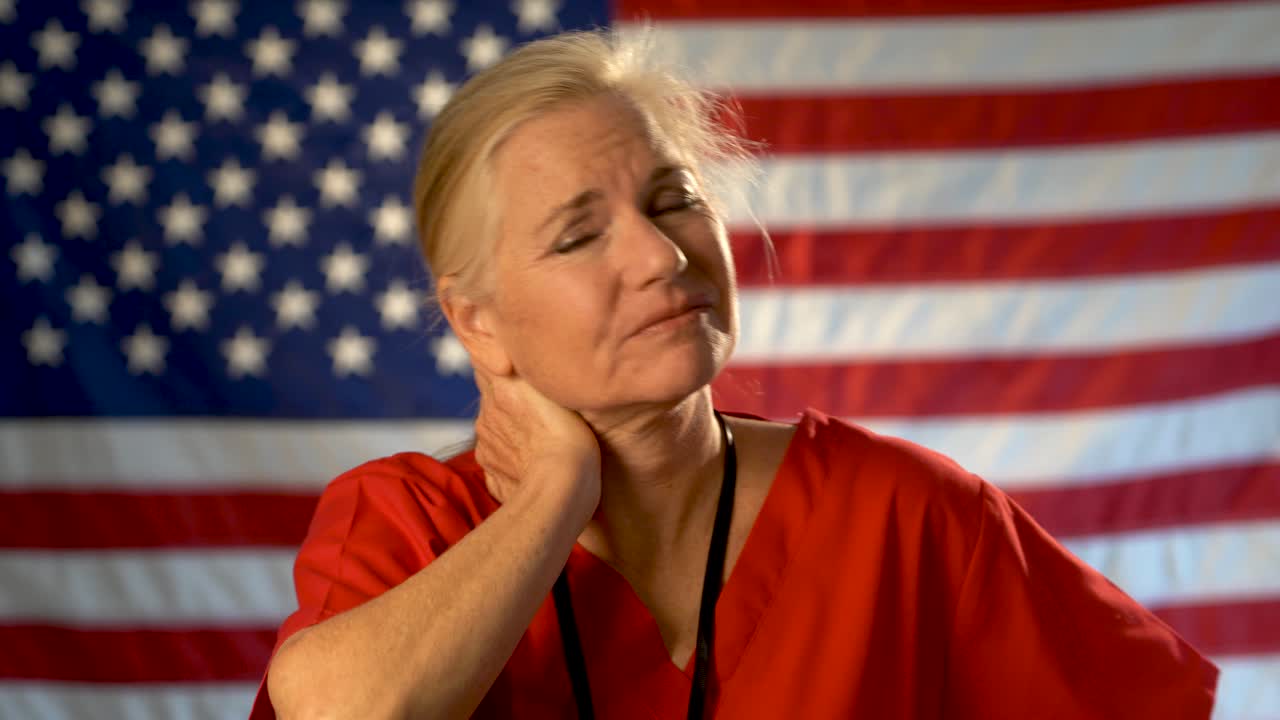 Medium tight portrait of nurse looking very worried and sad, hand on her forehead with American flag behind her