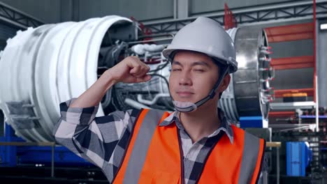 close up of asian male engineer with safety helmet flexing his bicep and smiling to camera while standing with airplane engine maintenance conducted, analytics checking the turbine