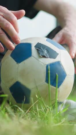a close-up of an elderly man seated on a grassy field, gently touching a soccer ball with both hands, with a blur background of trees