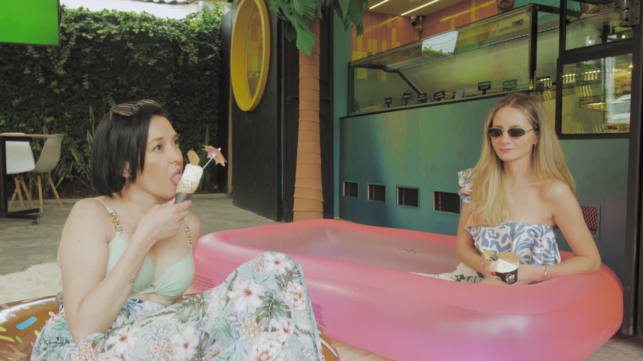 Two young women enjoy ice cream while relaxing in an inflatable pool in front of a modern ice cream parlor with tropical decor.