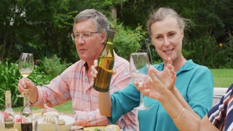 animation of diverse happy senior female and male friends eating lunch in garden, pouring wine