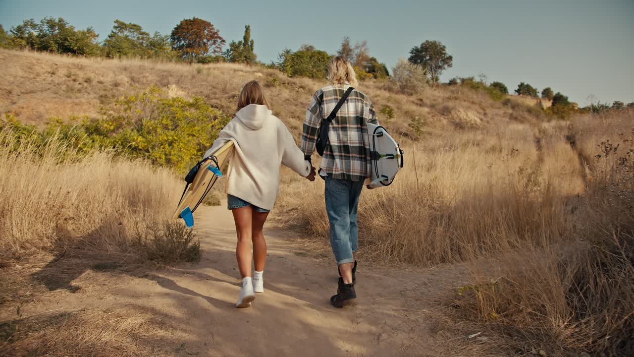 Rear view of a blond guy in a plaid shirt in jeans holding hands with his blonde girlfriend in a white sweatshirt. They are carrying surfboards and walking along a path with dried grass and small trees