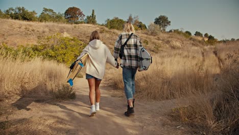 Rear-view-of-a-blond-guy-in-a-plaid-shirt-in-jeans-holding-hands-with-his-blonde-girlfriend-in-a-white-sweatshirt.-They-are-carrying-surfboards-and-walking-along-a-path-with-dried-grass-and-small-trees