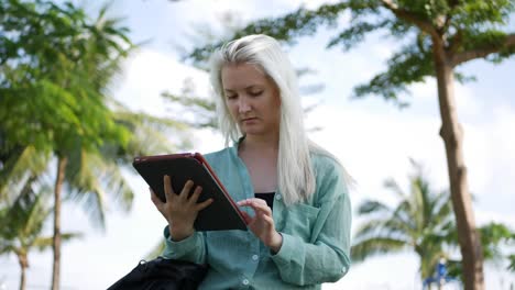 beautiful slim woman with long blonde hair in green shirt sits on the ground and using smartphone over background the park. girl on the square touching screen and smile