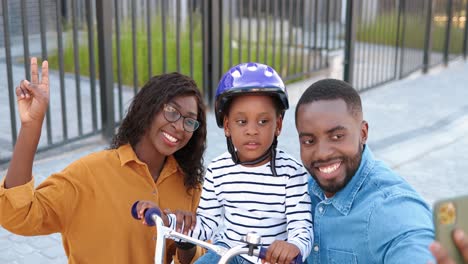 Porträt-Einer-Afroamerikanischen-Familie-Und-Einer-Süßen-Kleinen-Tochter-Im-Helm,-Die-Auf-Dem-Fahrrad-Sitzt-Und-Mit-Der-Smartphone-kamera-Lächelt-Und-Selfie-fotos-Macht