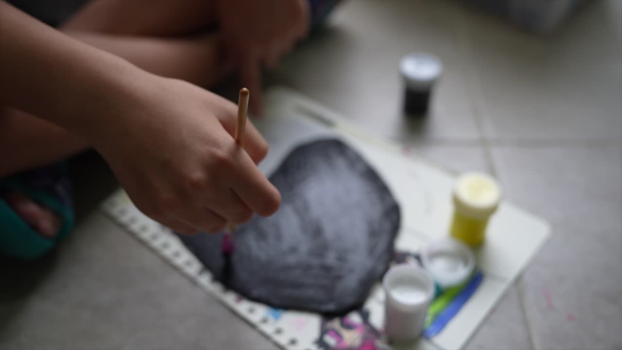 Close Up of Girl Painting in Natural Light. The scene is set in a well-lit indoor environment, where the focus is on the girl's hand holding a paintbrush as she applies paint to a sheet of paper
