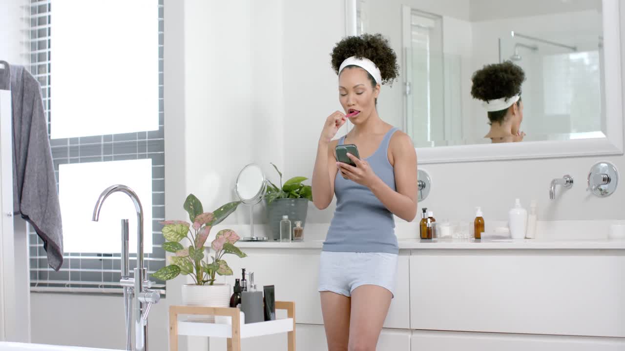 mujer biracial feliz cepillándose los dientes y usando un teléfono inteligente en el baño, cámara lenta