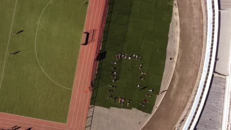 una grabación aérea giratoria dinámica de un grupo de estudiantes tirados en el césped del estadio deportivo mientras hacen ejercicios y movimientos de brazos